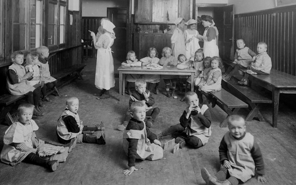 Vuxna och barn sitter på stolar och golv i ett rum tidigt 1900-tal. Foto.